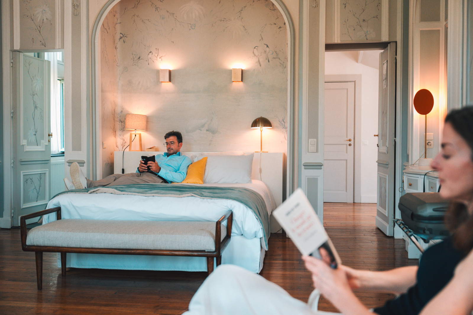 A man lies on a bed while his wife reads next to him at the Hôtel des Sorbiers in Hastière
