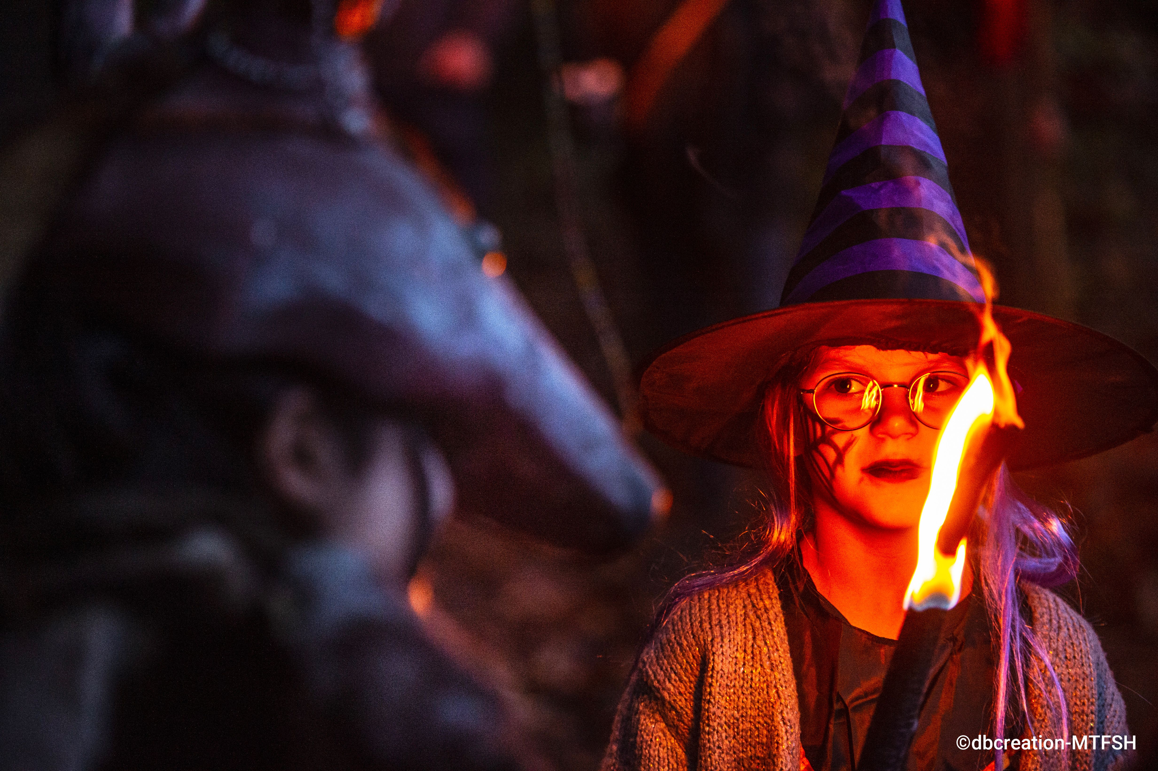Petite fille grimée et déguisée participant à une marche au flambeau dans les bois de Remagne et dont l'attention est attirée par une créature étrange