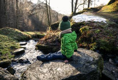 A child sits on a rock facing a river during a winter walk