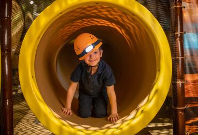 Kind mit gelbem Helm in einem Tunnel des Spielplatzes im Feriendorf Center Parcs Les Ardennes