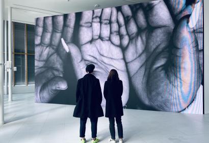 Deux personnes regardant une photo de mains exposée sur un pan de mur entier.