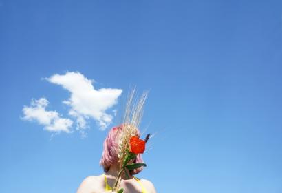 Femme dont le visage est caché par un bouquet d'épis de blé et de coquelicots, avec pour fond un ciel bleu.