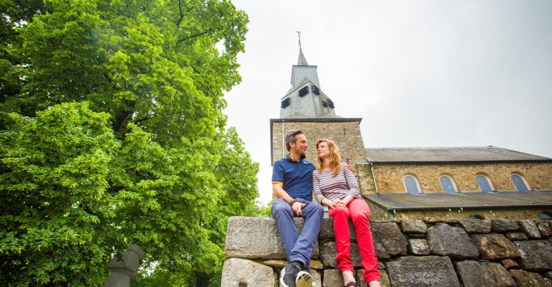 Couple sitting on a small stone wall by the Romanesque Saint Etienne church in Waha